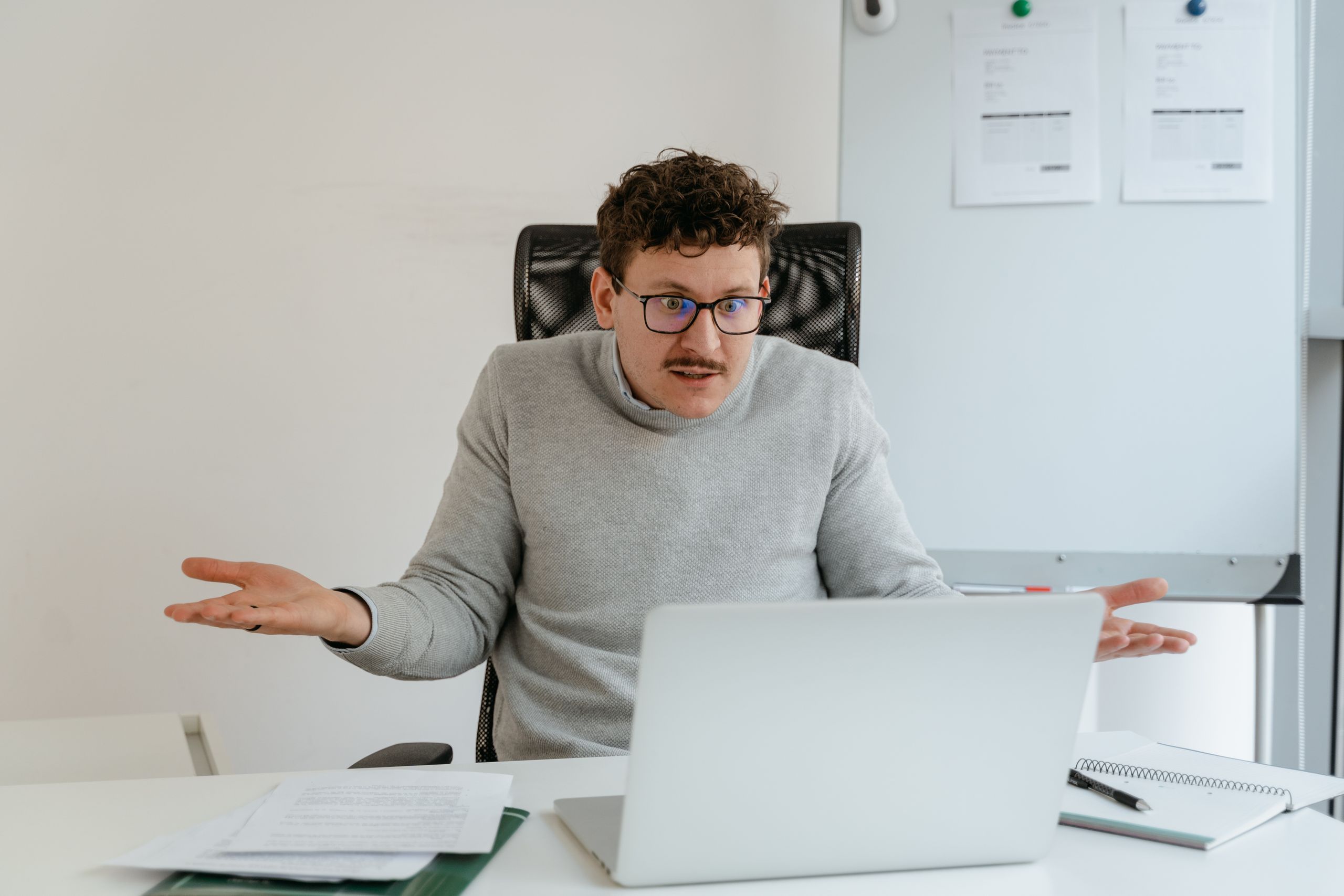 A man in a gray sweater and glasses gesturing at his laptop screen during a video call in an office setting.
