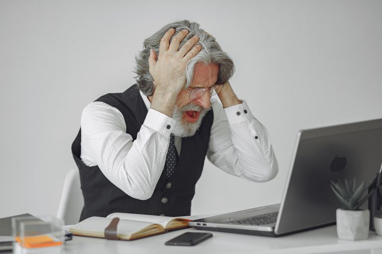 Senior businessman sitting at desk, expressing frustration with his laptop in a professional office setting.