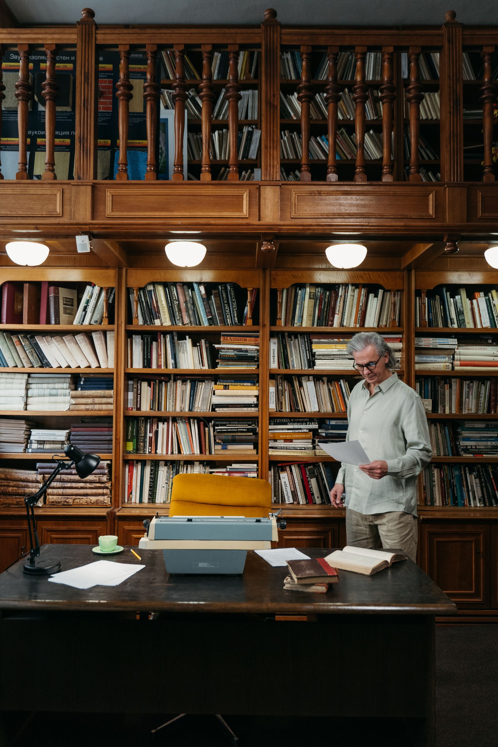Senior man reading in a library with an old typewriter and shelves full of books.